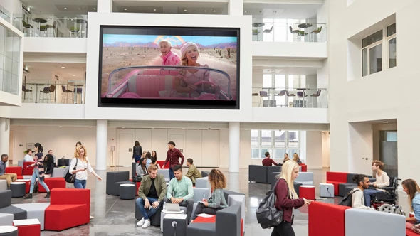 The film Barbie is being played on a bit TV screen in the background of a hall area at a university